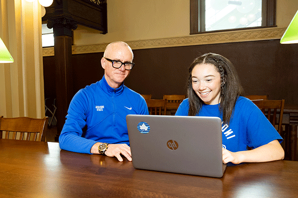 A student and an instructor sit together at a wooden table, smiling while looking at an HP laptop. The student wears a blue 'Indiana State' shirt, and the instructor wears a matching blue Indiana State jacket. The setting appears to be a study area with warm lighting and a classic interior design.